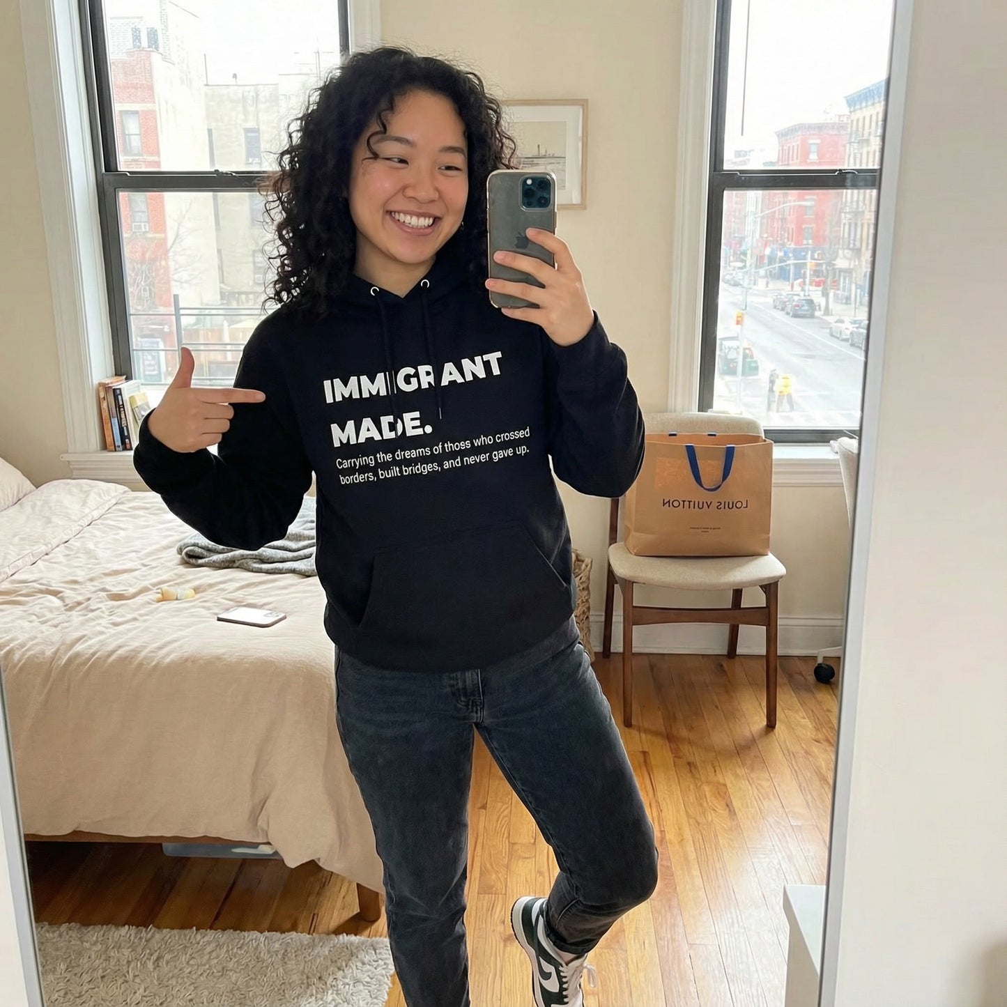 Person wearing a hoodie with 'IMMIGRANT MADE' text, taking a mirror selfie in a room.